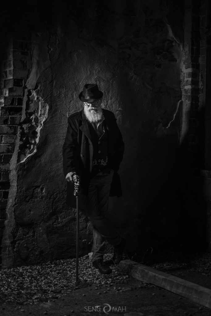 A moody black and white picture of a bearded man dressed in a hat and jacket, holding a walking stick and standing next to an old wall.