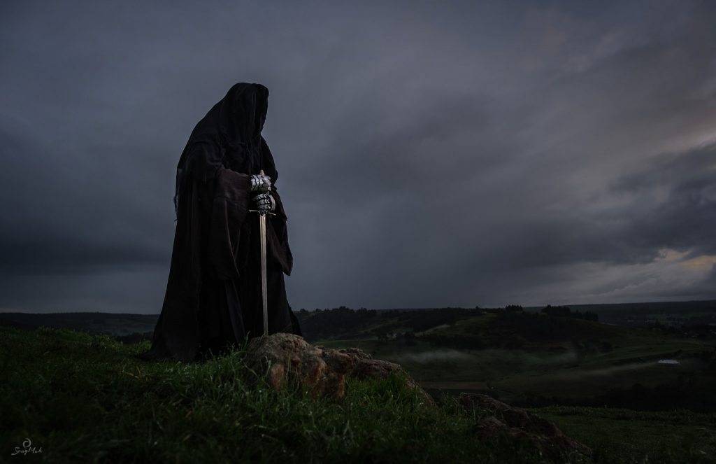 A man dressed as a Nazgul or Ring Wraith, completely covered in a hooded cloak and holding a sword, stands on a rock.