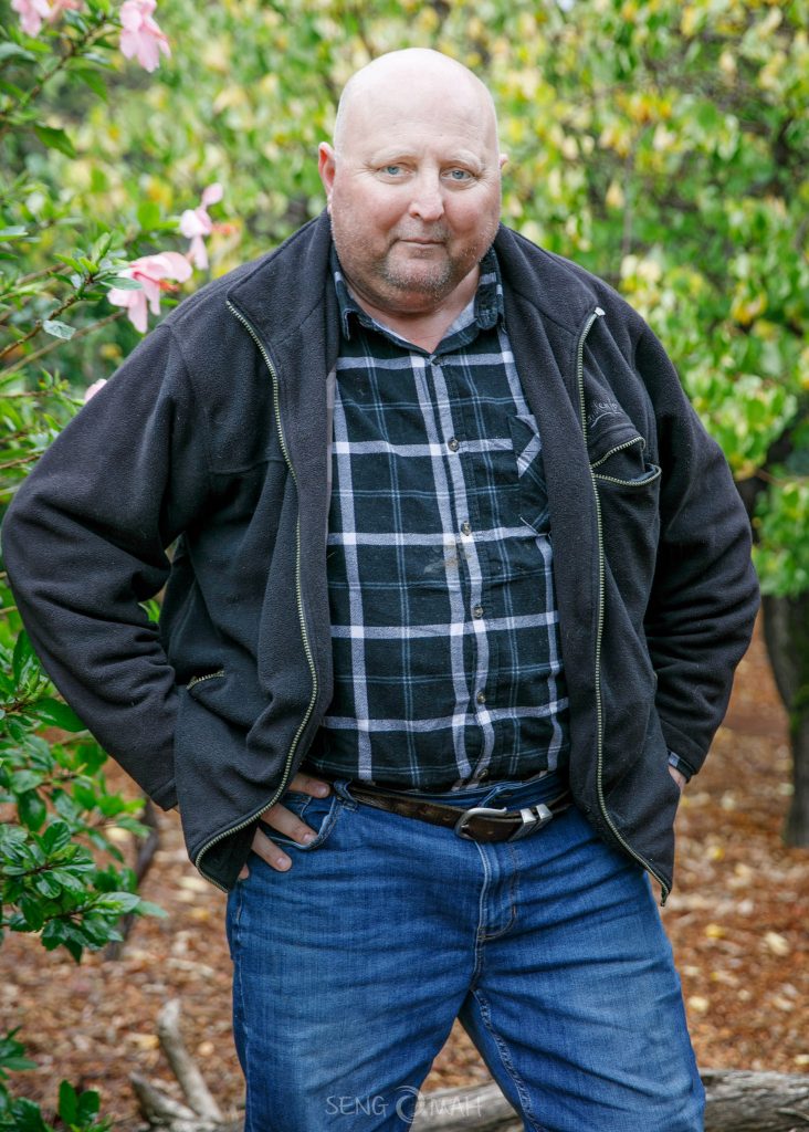 A portrait of Steve Marijanich aka Marra taken in his garden. Steve is wearing a flannel shirt, a jacket and jeans.