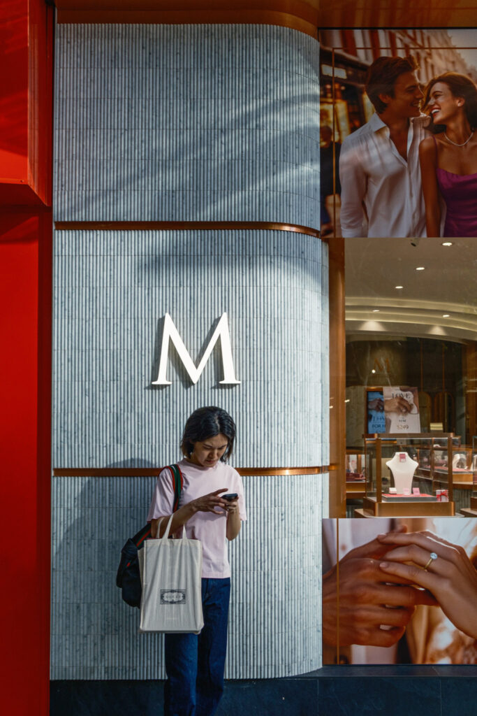 Street photograph of a girl checking her smartphone in front of an M sign and shop posters.
